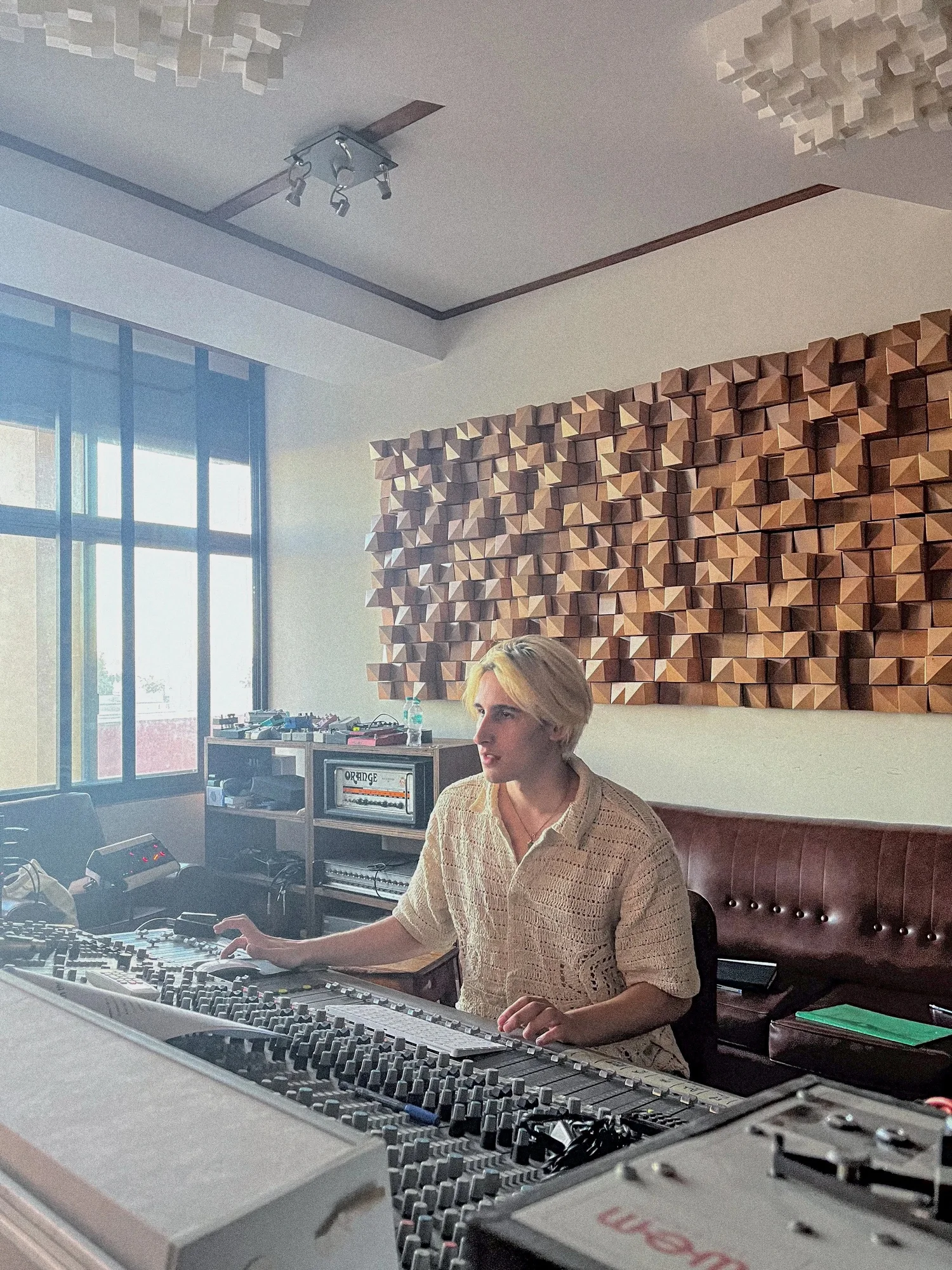 Lucas Mirovski sitting at a mixing console in a studio with acoustic panels on the wall and large windows letting in natural light.